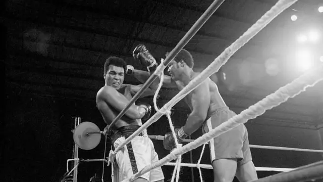 George Foreman punches Muhammad Ali by the ropes at the edge of the ring. The image is in black and white