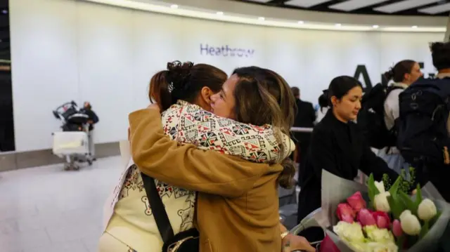 Two women share a hug with one holding a trolley with a bunch of flowers. Other passengers can be seen behind them