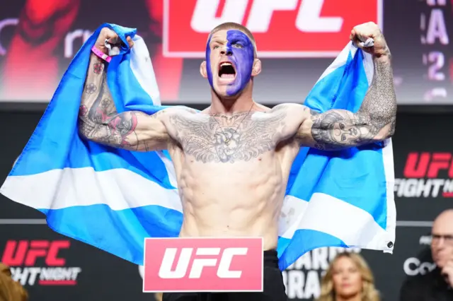 Chris Duncan holds up the Scotland flag as he stands on the scales at a UFC weigh-in