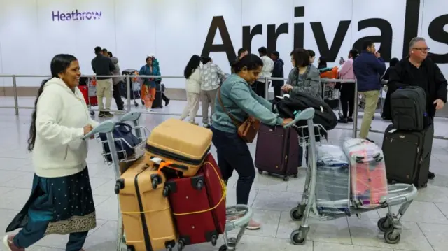 A number of people are pushing luggage on trolleys with a large 'Arrivals' sign in the background