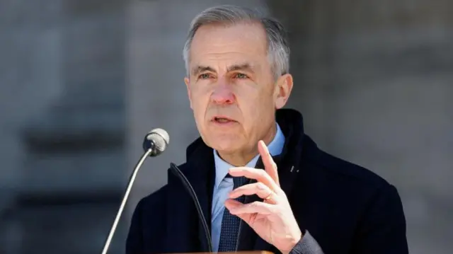 Carney speaking into a microphone, holding one hand up in front of his chest point upwards