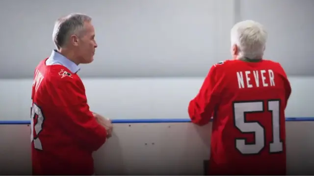 Mark Carney and Mike Myers wear hockey jerseys, with Myers' back showing "NEVER 51"