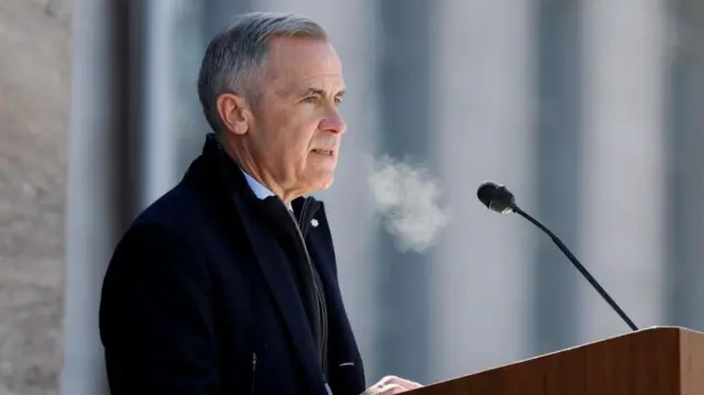 Carney speaking into a microphone, from side on. You can see his breath, meaning it must be cold