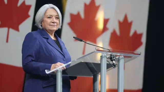 Woman in purple suits stands at podium with Canadian flags in background