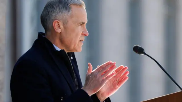 From side on, Carney speaks at a lectern with a small black press microphone. He holds both hands up out in front of his chest with palms facing each other