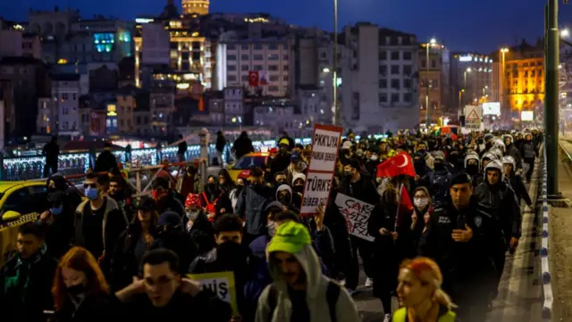 A large group of protesters make their way across a bridge. Some are holding signs written in Turkish, a few are holding Turkish flags.