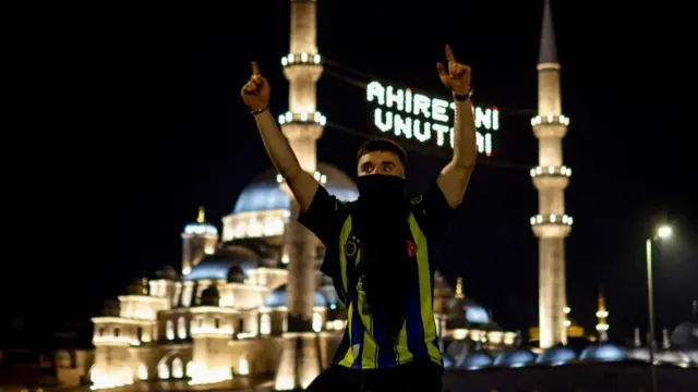 A man in a Fenerbache shirt and mask over part of his face with his arms raised. There is a mosque, lit up in the night sky, behind him.