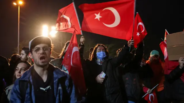 Protesters holding Turkish flags. A woman in a blue face mask is carrying an iPhone.