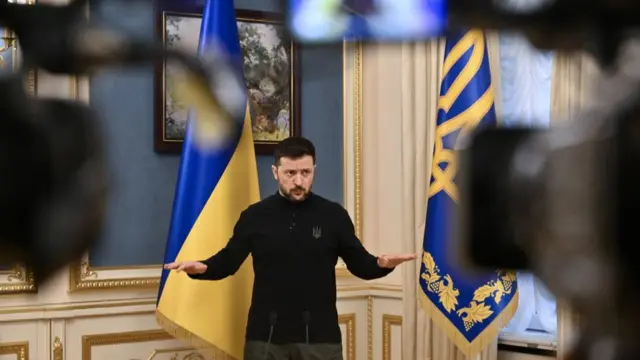 Volodymyr Zelensky stands with his hands outwards in front of two Ukrainian flags while speaking to reporters.