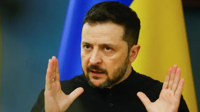 Volodymyr Zelensky holds up his hands against the backdrop of a Ukrainian flag.