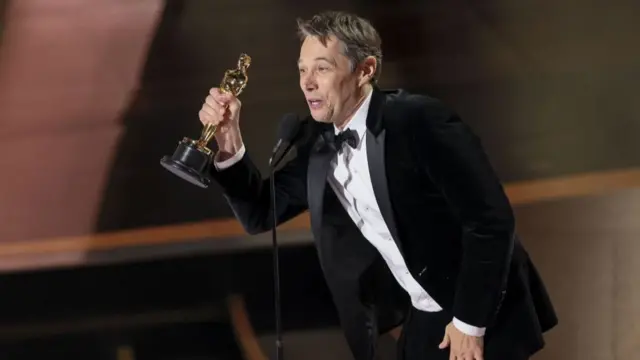 Sean Baker holding one of his four Oscars wearing a black velvet suit and bow tie