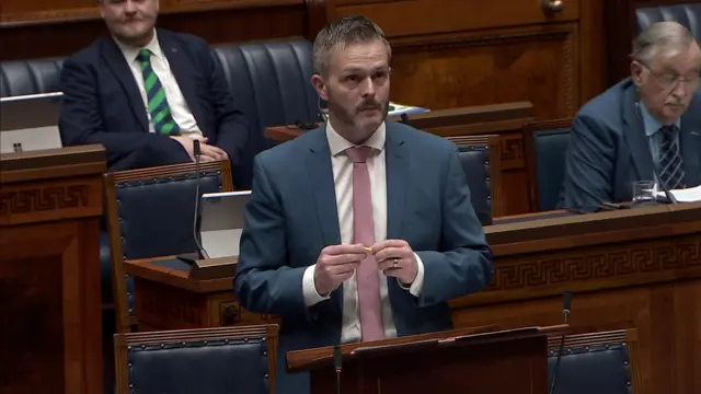 Robbie Butler, a man with short, greying hair and a beard, is addressing the NI Assembly.  He is wearing a blue suit, white shirt and pink tie.