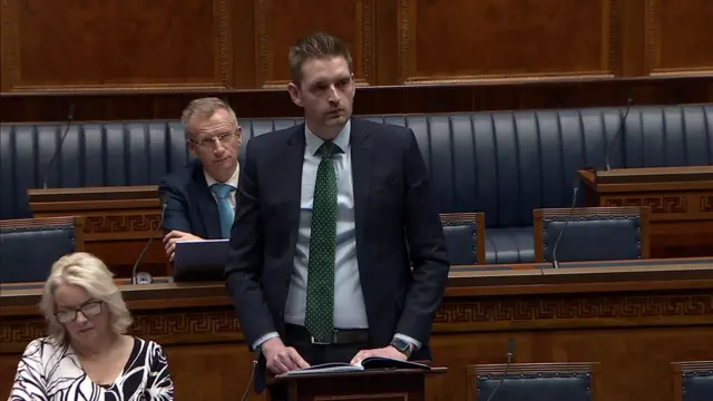 Phillip Brett speaking in the assembly. He is wearing a dark suit, blue shirt and green tie.