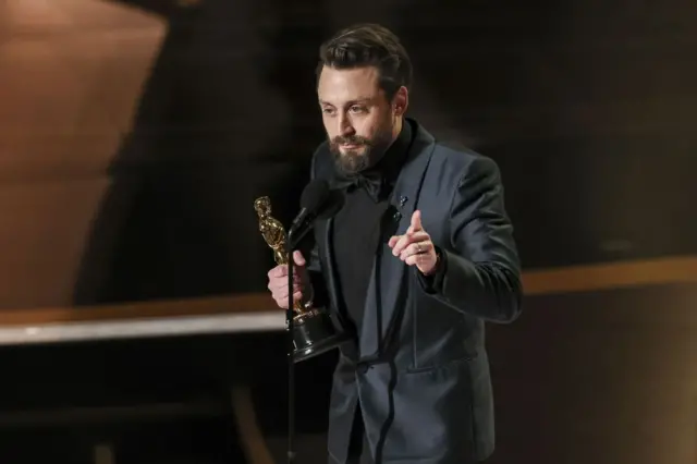 Kieran Culkin holding his Oscar in a black suit, shirt and bow tie