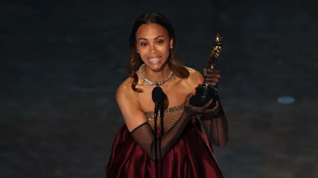 Zoe Saldaña holding her oscar on the stage in a burgundy dress and black sheer gloves