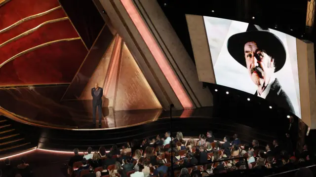 Morgan Freeman on stage at the Oscars with an image of the late Gene Hackman appearing on a board behind him