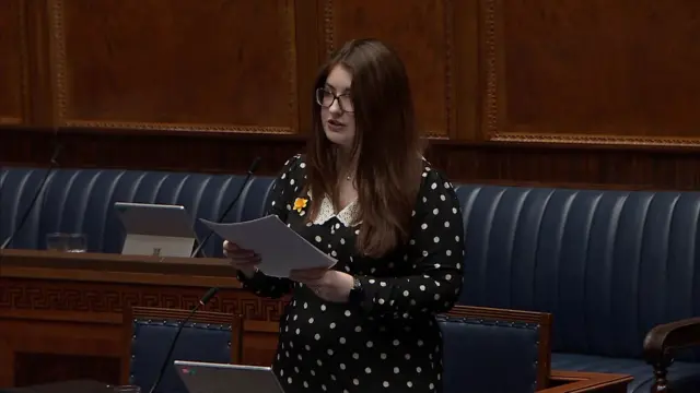 Connie Egan addressing the NI Assembly.  She has long, straight brown hair and is wearing dark-rimmed glasses and a black and white polka dot dress.