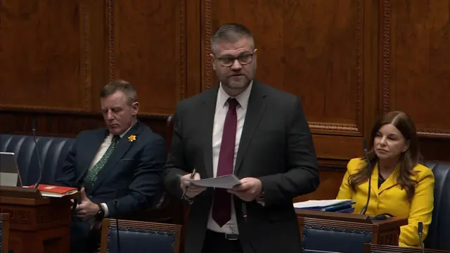 Colin McGrath addressing the NI Assembly.  He has short grey hair, glasses and is wearing a grey suit with a maroon tie.