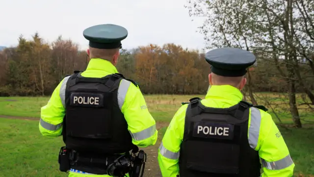 Two uniformed police officers walk side by side with their backs to the camera. The are wearing black armoured vest and yellow high-vis coats.  They are walking towards a grassed, wooded area.