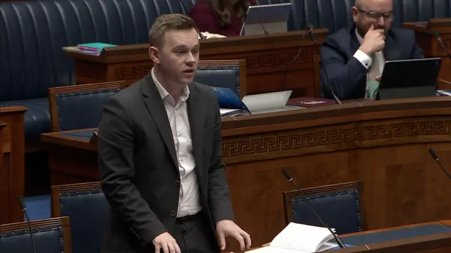 Eóin Tennyson addressing the assembly.  He has short, fair hair and is weaing a grey suit with a pale, open-neck shirt.