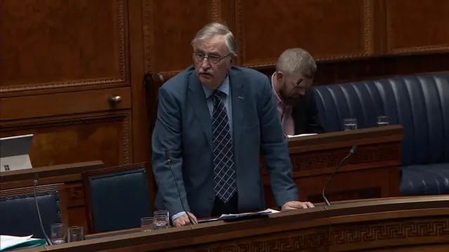 Alan Chambers addressing the NI Assembly.  He has grey hair and a grey moustache.  He is wearing a blue suit with a navy patterned tie.