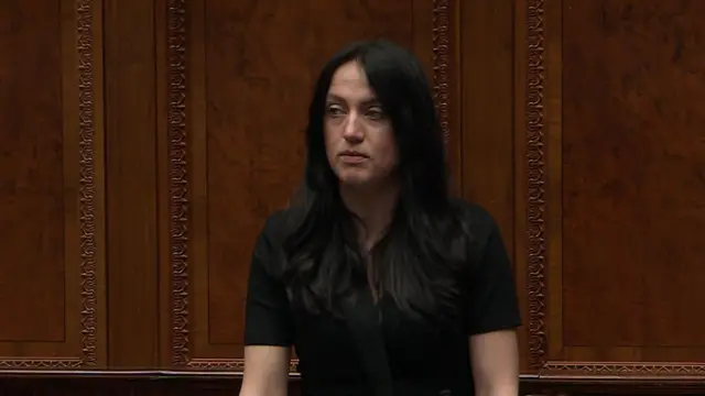 A woman with long black hair is looking off to the side. She is wearing a black shirt and trousers.