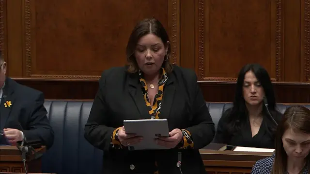 Deirdre Hargey, has short brown hair and is wearing a orange/yellow top with black and white patterns, and a black jacket. She is holding a paper booklet and reading from it.