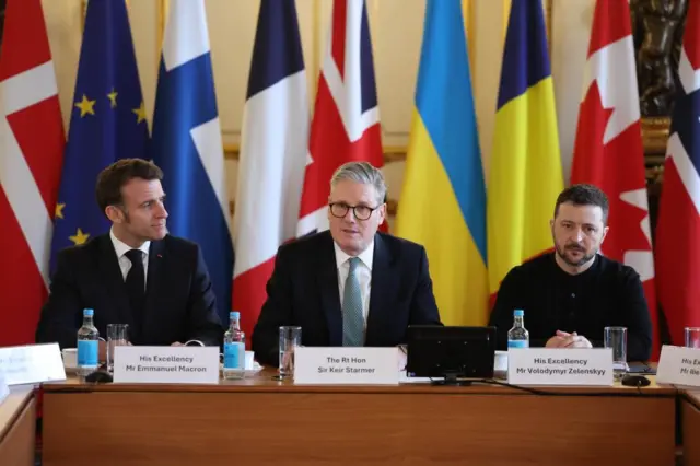 Macron, Starmer and Zelensky sit at a table with their flags draped behind