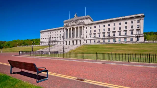 A large white stone building sits on top of a hill. The sky is blue and in the foreground is green grass, a red road and a wooden bench facing the building.