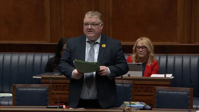 Declan McAleer addressing the NI Assembly.  He has short, greying blonde hair and is wearing a navy suit, blue shirt and a patterned blue tie.