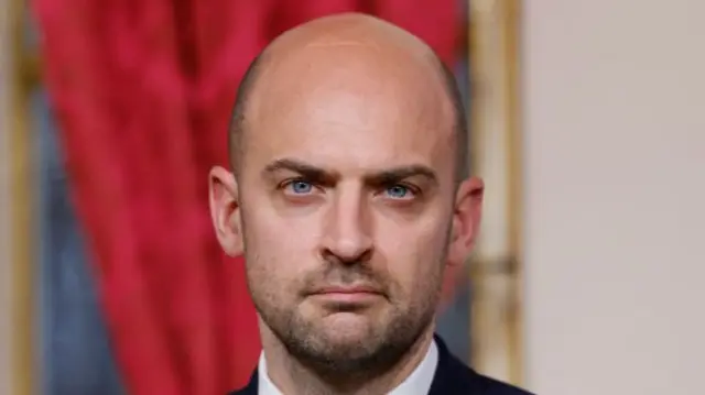 Barrot is staring at the camera. In the background, there is a red curtain to the left and a plain beige wall to the right.