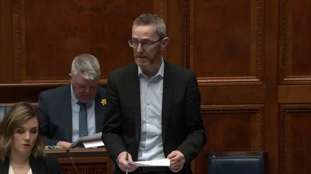 Philip McGuigan standing in the assembly wearing a dark suit and white shirt. He is wearing glasses and has a red and grey beard and hair.