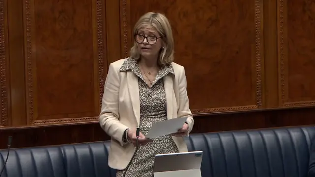 Ulster Unionist Diana Armstrong addressing the assembly. She as shoulder-length blonde hair and black-framed glassed.   She is wearing a cream suit jacket over a taupe patterned dress.