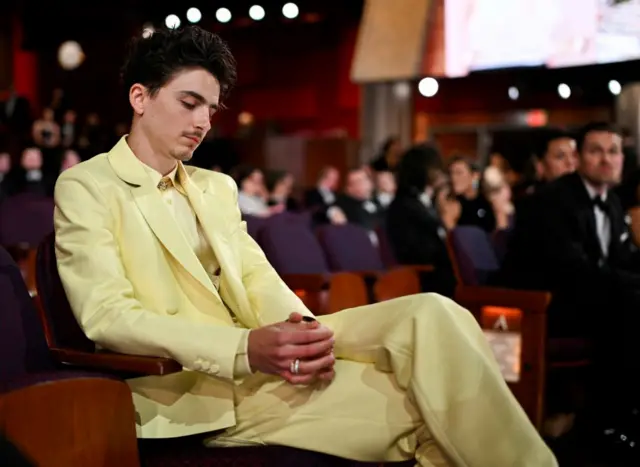 Timothee Chalamet sat looking pensive with his hands clasped, wearing a yellow suit