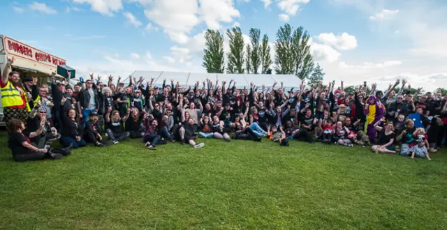 A very large group of festival goers are gathered around a section of grass, posed for a group photograph.