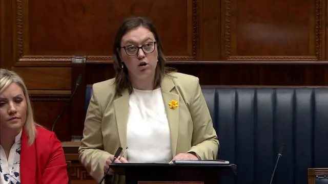 Deborah Erskine who has medium brown hair, is wearing a white top and off yellow jacket and black framed glasses.