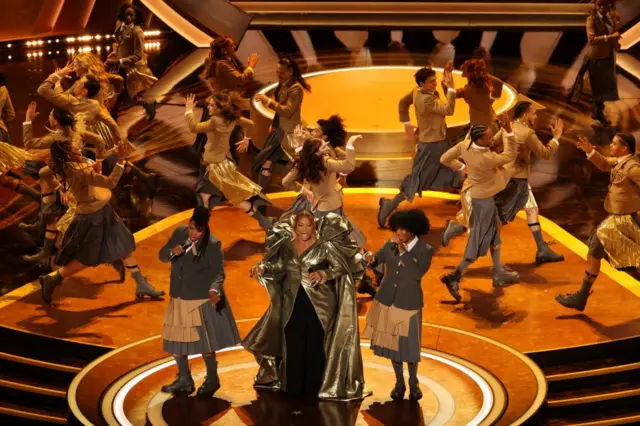 Queen Latifah wearing a golden cape surrounded by dancers on the Oscars stage