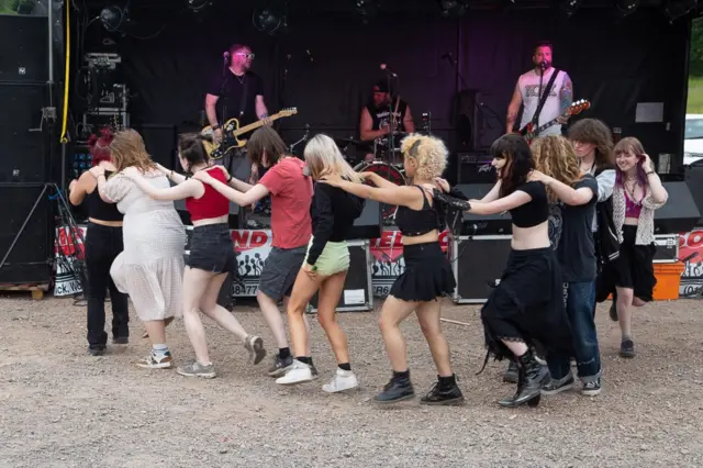 A band plays music on a small stage whilst 10 people do the conga line in front.