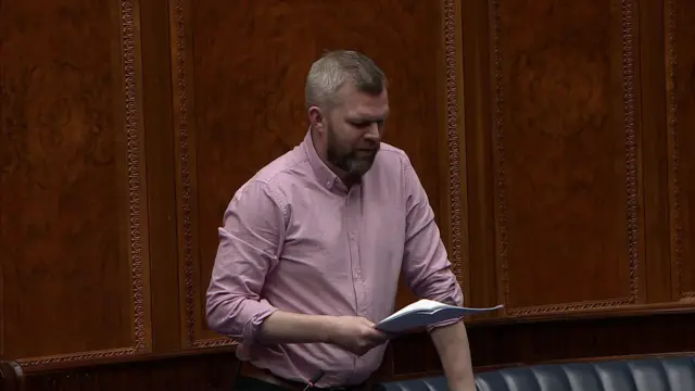 Gerry Carroll addressing the NI Assembly. He has short grey hair and a beard and is wearing a pink open-neck shirt.