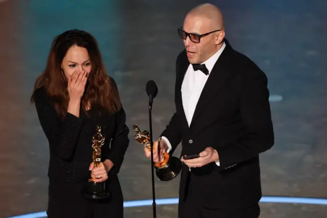 Shirin Sohani with her hand covering her mouth in surprise and Hossein Molayemi holding their Oscars