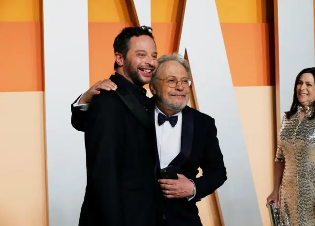 Billy Crystal with his arm around Nick Kroll, both in black suits