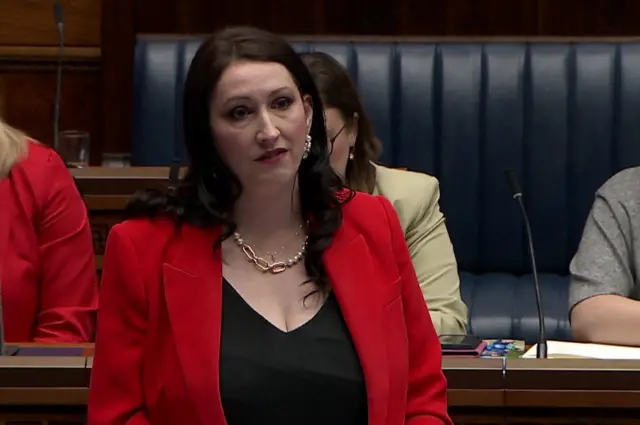 Emma Little-Pengelly in the assembly. She has long black hair and is wearing a gold necklace and earrings. She is wearing a red jacket and black top.