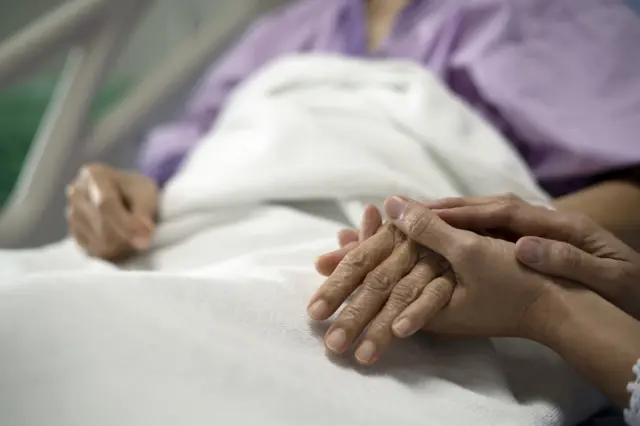 An elderly patient is lying in a hospital bed, a white sheet is pulled up around her chest. A woman sitting beside her bed is holding her hand.