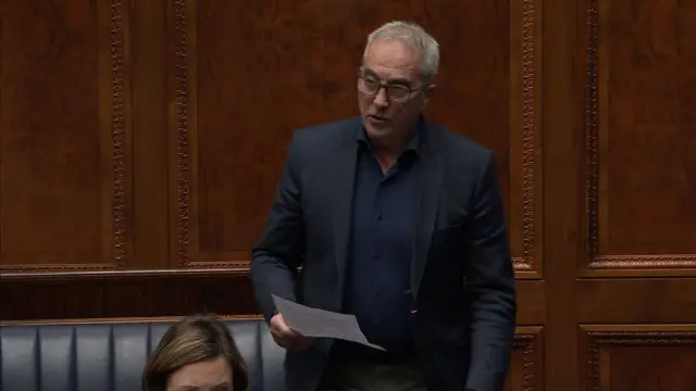 Pat Sheehan who has grey short hair, is wearing a navy shirt and dark suit jacket. He has black framed glasses on and is holding a piece of paper as he speaks.