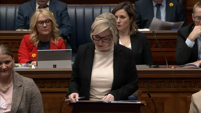 Mchelle O'Neill with blonde hair and a side fringe. Her hair is up. She is wearing a cream jumper and black jacket standing in the assembly.