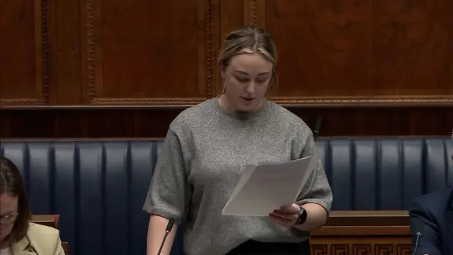 Cheryl Brownlee has her blonde hair tied up and is wearing a grey short sleeved jumper. she is holding a statement in front of her and is reading it out at the assembly.