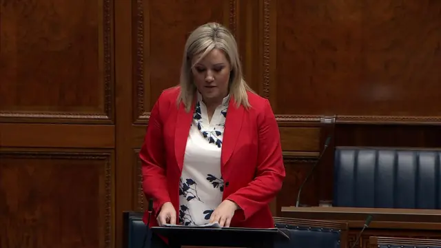 Diane has medium length blonde hair. She is wearing a white and black floral patterned shirt and a red jacket.