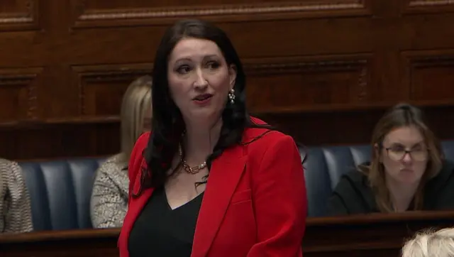 Emma Little-Pengelly in the assembly. She has long black hair and is wearing a gold necklace and earrings. She is wearing a red jacket and black top.