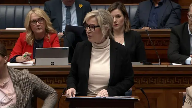 Michelle O'Neill addressing the Stormont chamber. She has blonde hair tied back and is wearing a black suit jacket and a cream turtleneck jumper.