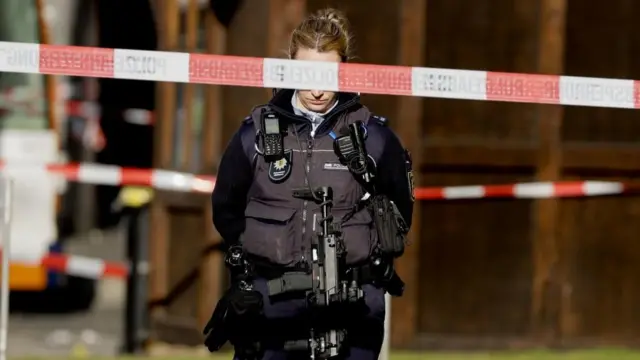 Police tape can be seen in the foreground, with a heavily armed police officer in the background.